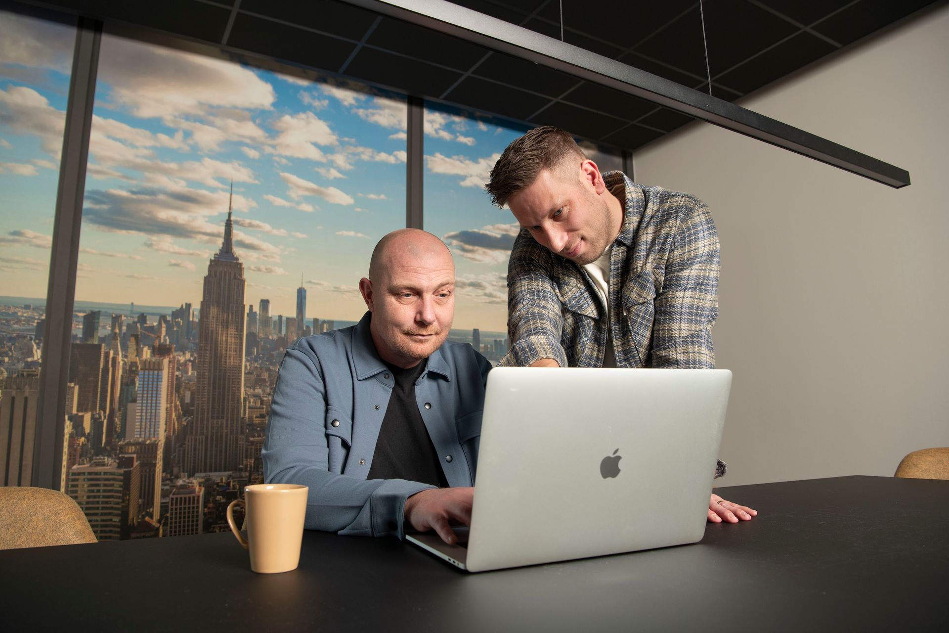 A person in a beige sweater sits at a wooden table, looking at a smartphone while a laptop is open in front of them.
