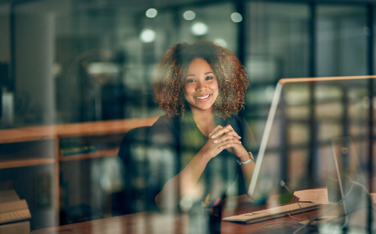 A smiling professional sits at a desk with clasped hands, viewed through a glass partition in a blurred office setting.