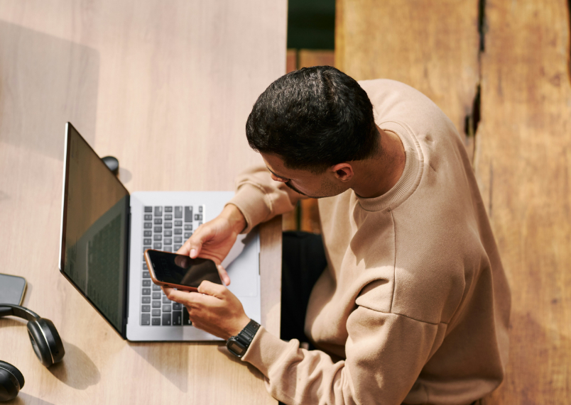 A person in a beige sweater sits at a wooden table, looking at a smartphone while a laptop is open in front of them.
