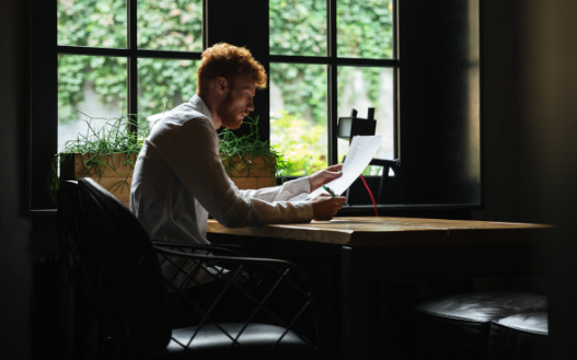 A person with reddish hair sits at a wooden table by a window, reading a document in a dimly lit, cozy indoor space.