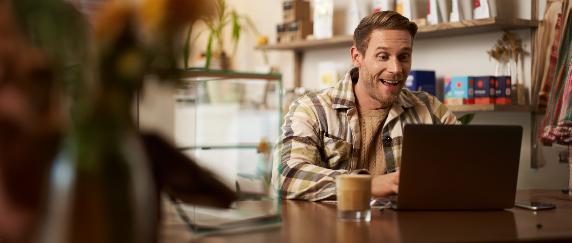 A person in a plaid shirt sits at a wooden table in a cafe, looking surprised while working on a laptop.