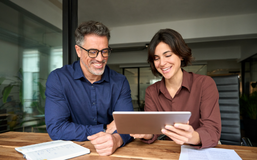 Two professionals in a modern office review information together on a tablet while sitting at a wooden desk.