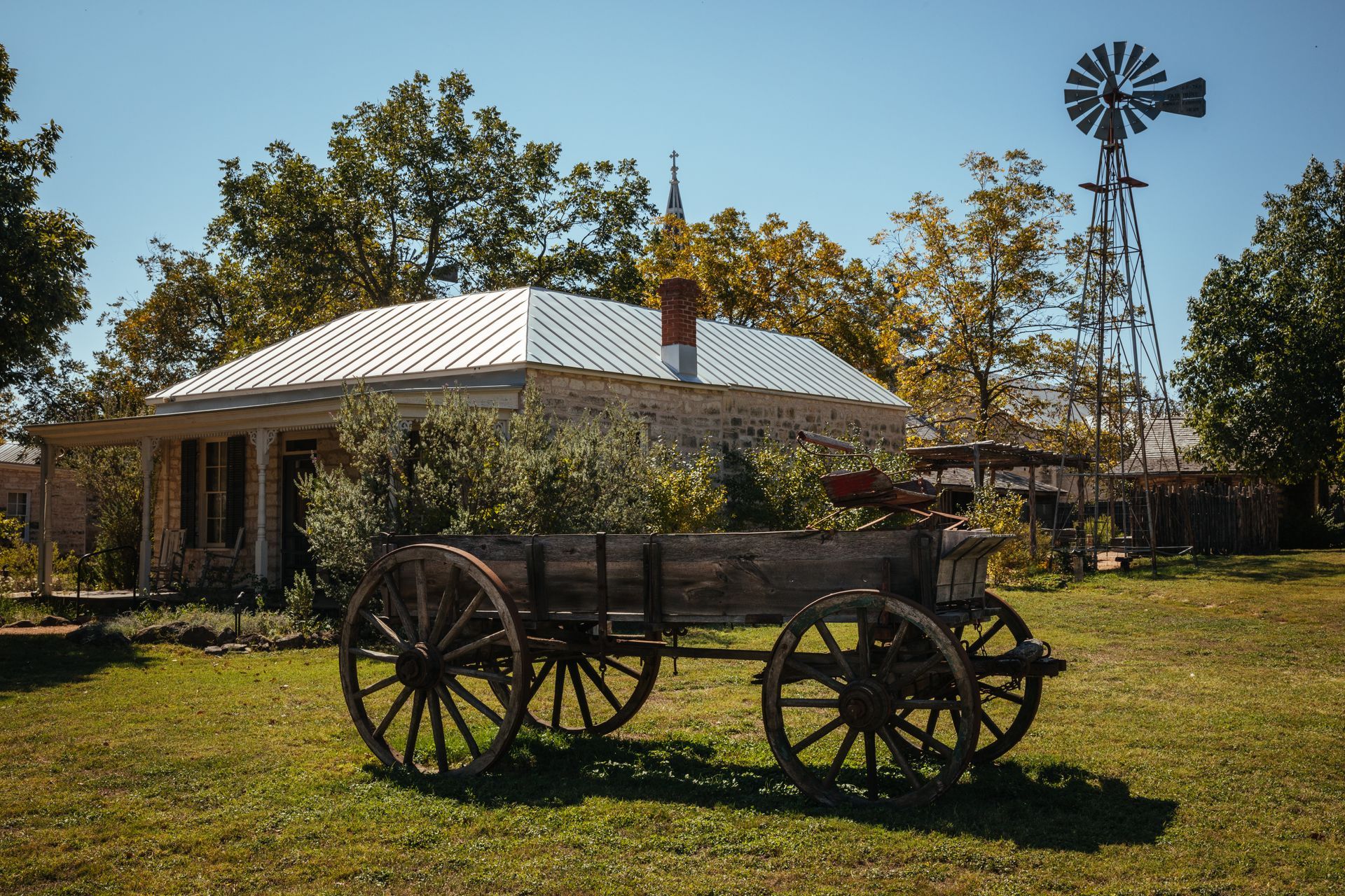 A wooden wagon is parked in a grassy field in front of a house and windmill.