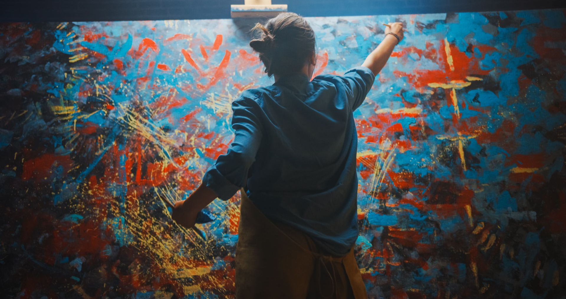 Person painting abstract art on a large canvas, reaching with both arms, splashes of blue, red, and yellow paint.