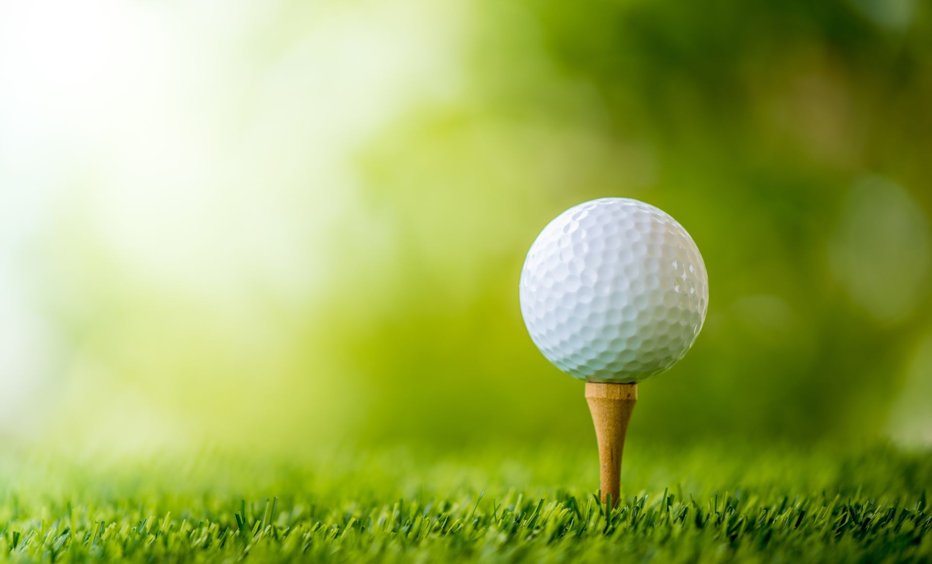 A white golf ball sits on a wooden tee embedded in vibrant green grass against a softly blurred, sunny background.
