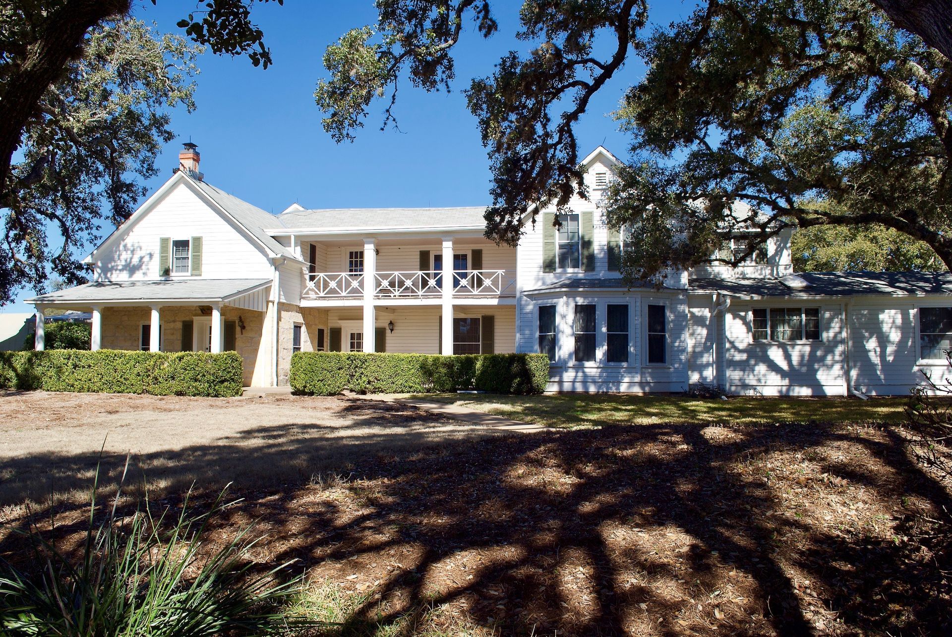 White two-story house with wrap-around porch, balcony, and mature trees casting shadows.