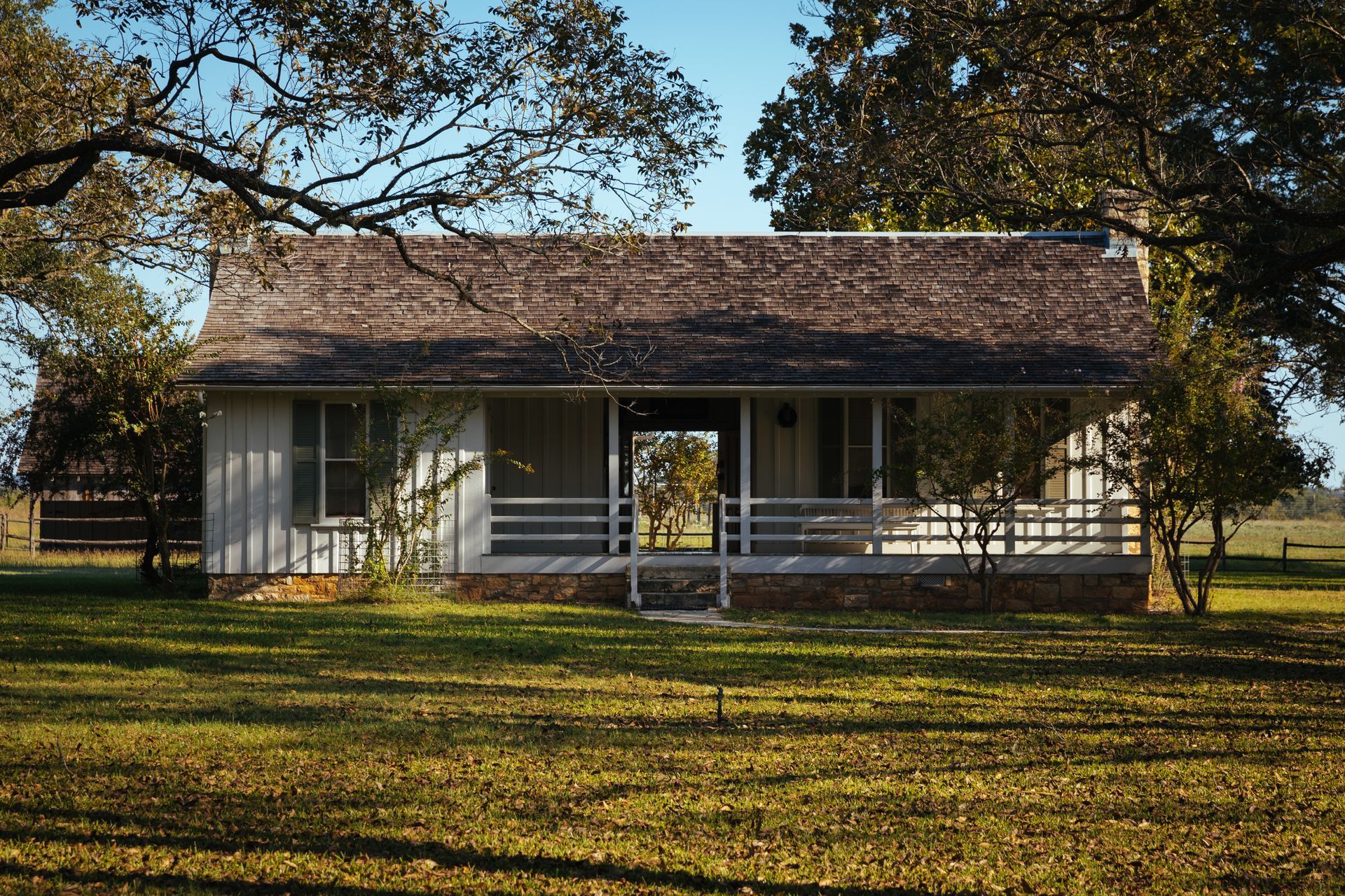 Small, white farmhouse with porch under a brown shingled roof, set in a grassy yard under trees.