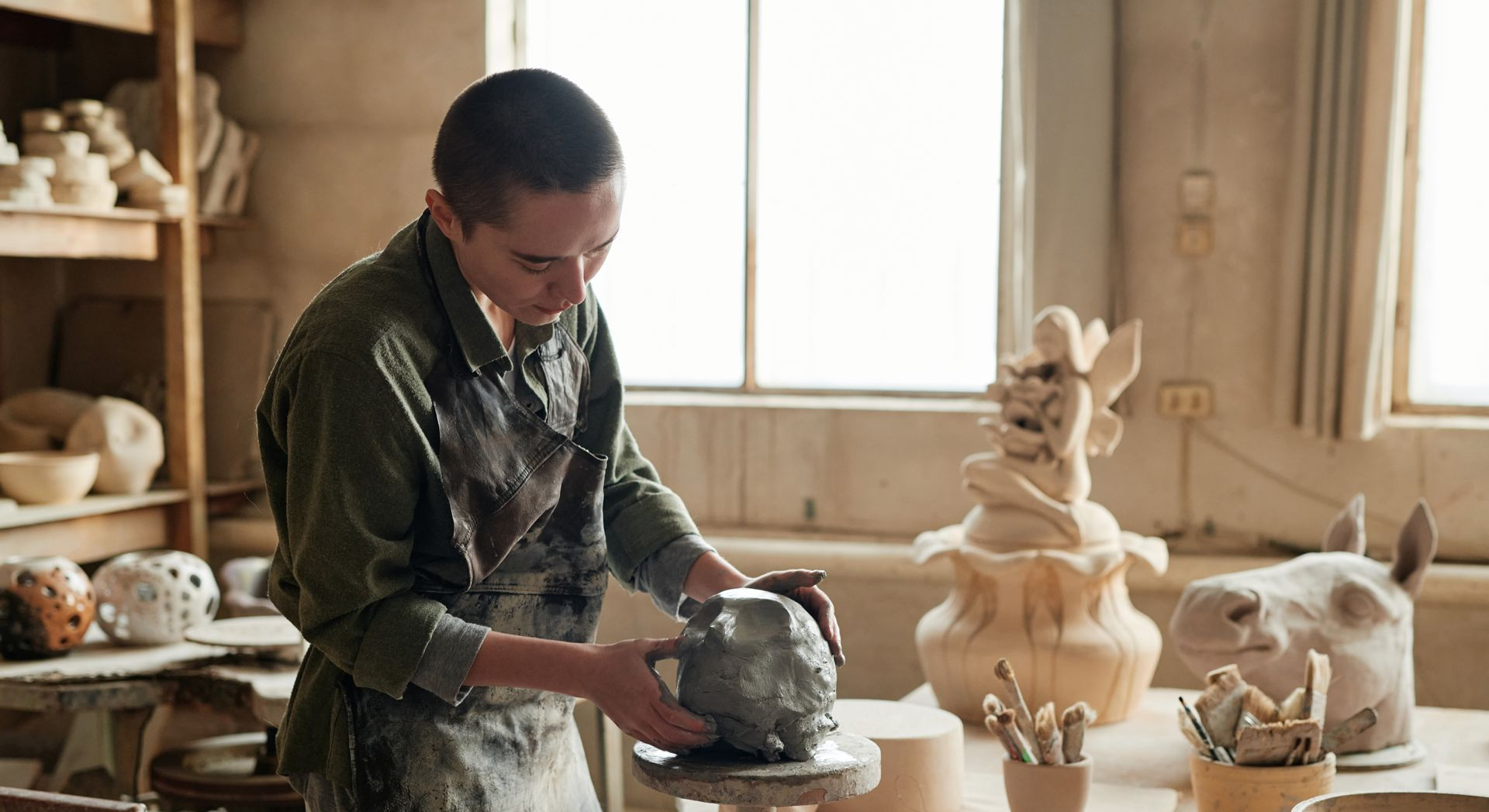 A person in a workshop sculpts a clay form on a pottery wheel.