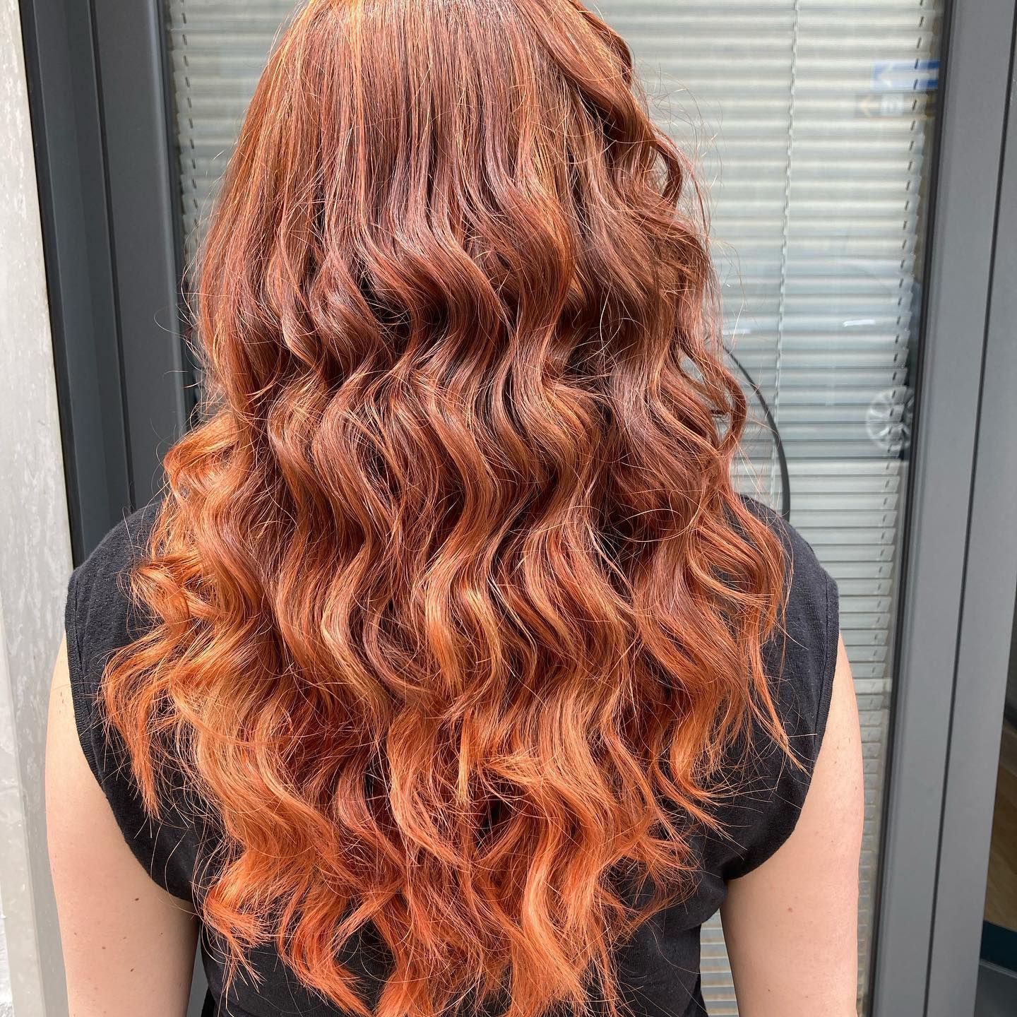 Donna con lunghi capelli castani ondulati. Vista posteriore, indossa un top nero, davanti a una finestra.