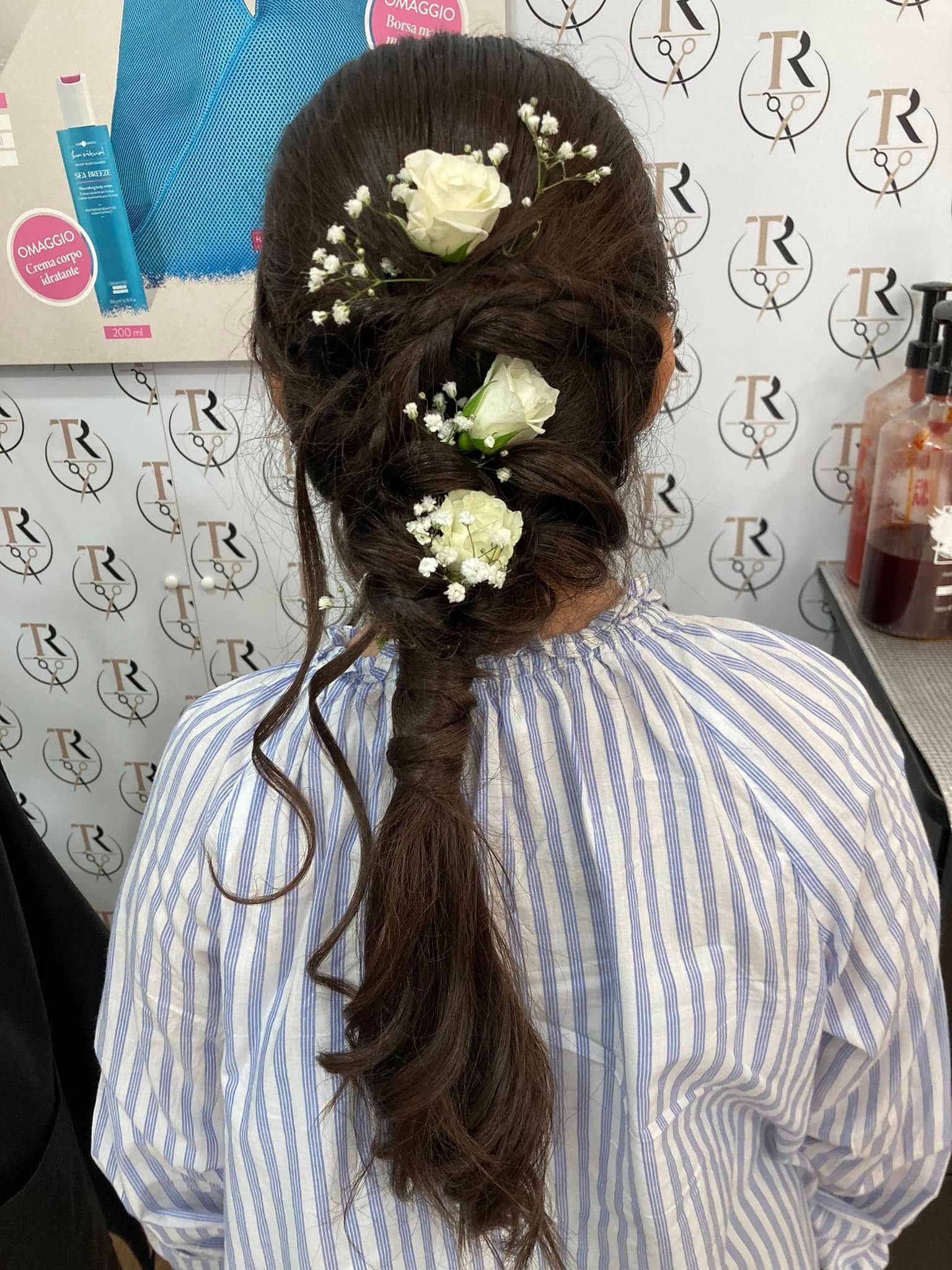 Schiena di donna con elaborata acconciatura castano scuro: tre mazzi di fiori e treccia, camicia a righe blu e bianche.