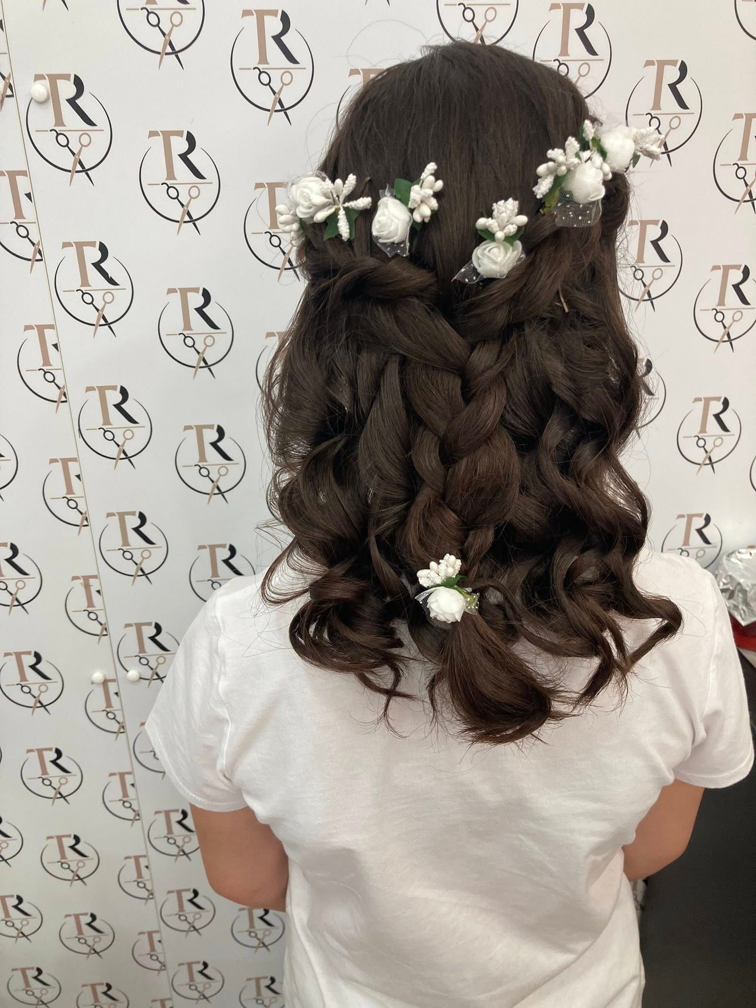 Donna con capelli scuri e ondulati, acconciati con una corona di fiori e trecce, che indossa una camicia bianca.