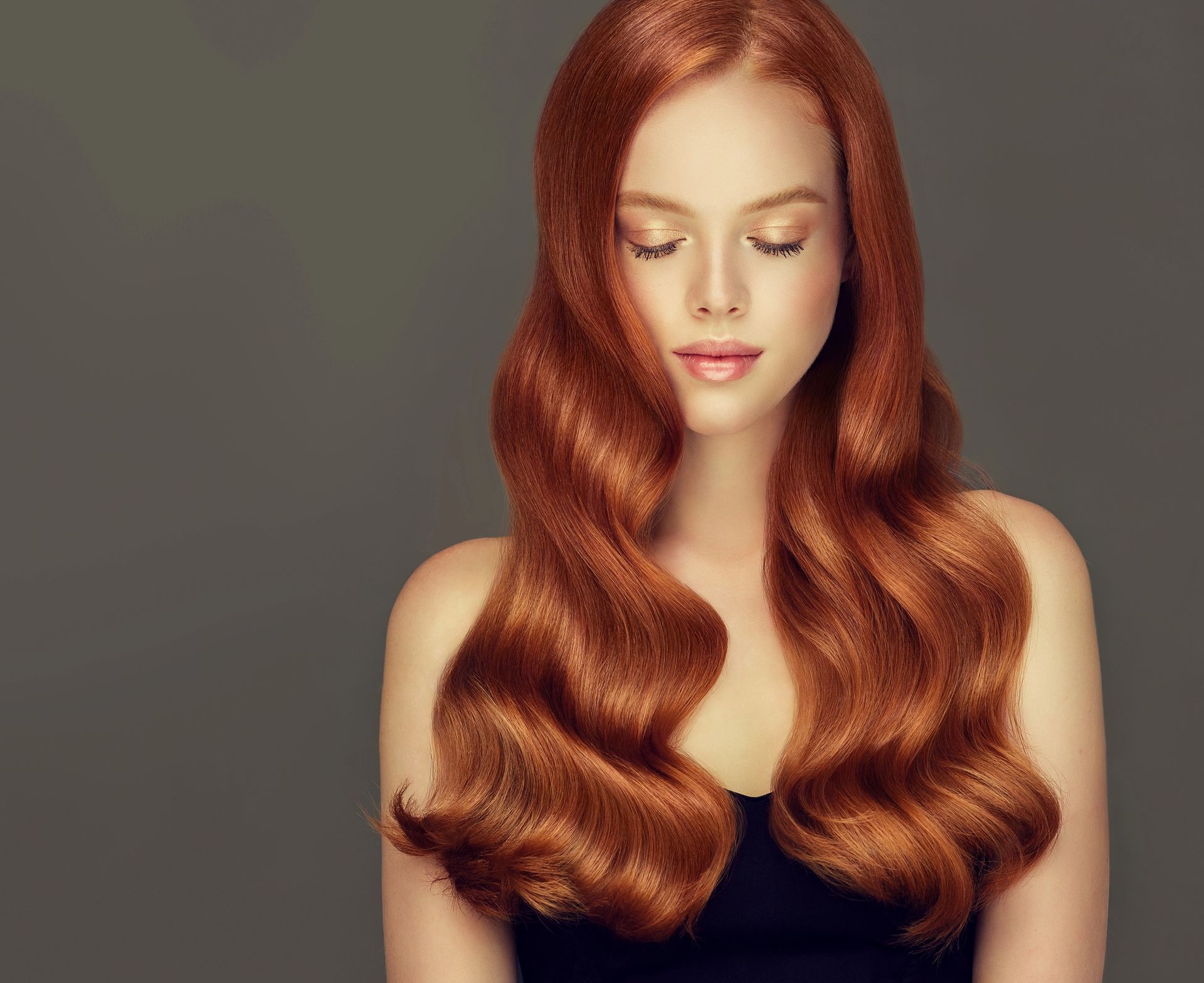 Donna con lunghi capelli castani ondulati, sguardo rivolto verso il basso e occhi chiusi, top nero, sfondo grigio.