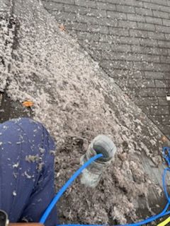 Person on a roof cleaning debris. Gray shingles covered in lint and dust. Blue hose connected to a cleaning tool.