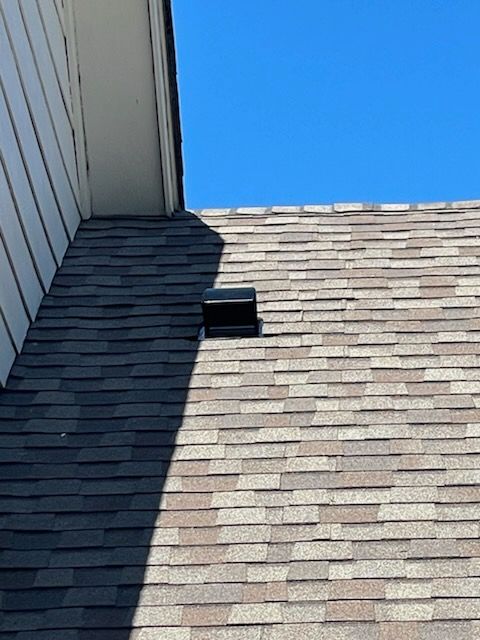 Roof with shingles, vent, and part of a white wall under a blue sky.