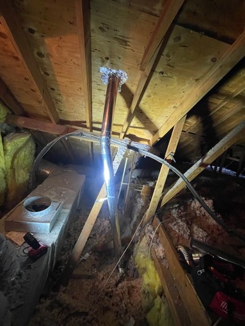 Copper pipe venting in an attic. Pipe bends, connected to a metal box, amidst insulation and wooden beams.