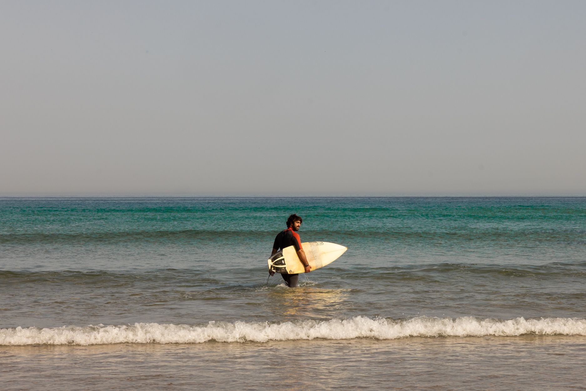 Un surfista sostiene una tabla de surf blanca en aguas poco profundas del océano, con olas tranquilas y un horizonte despejado.