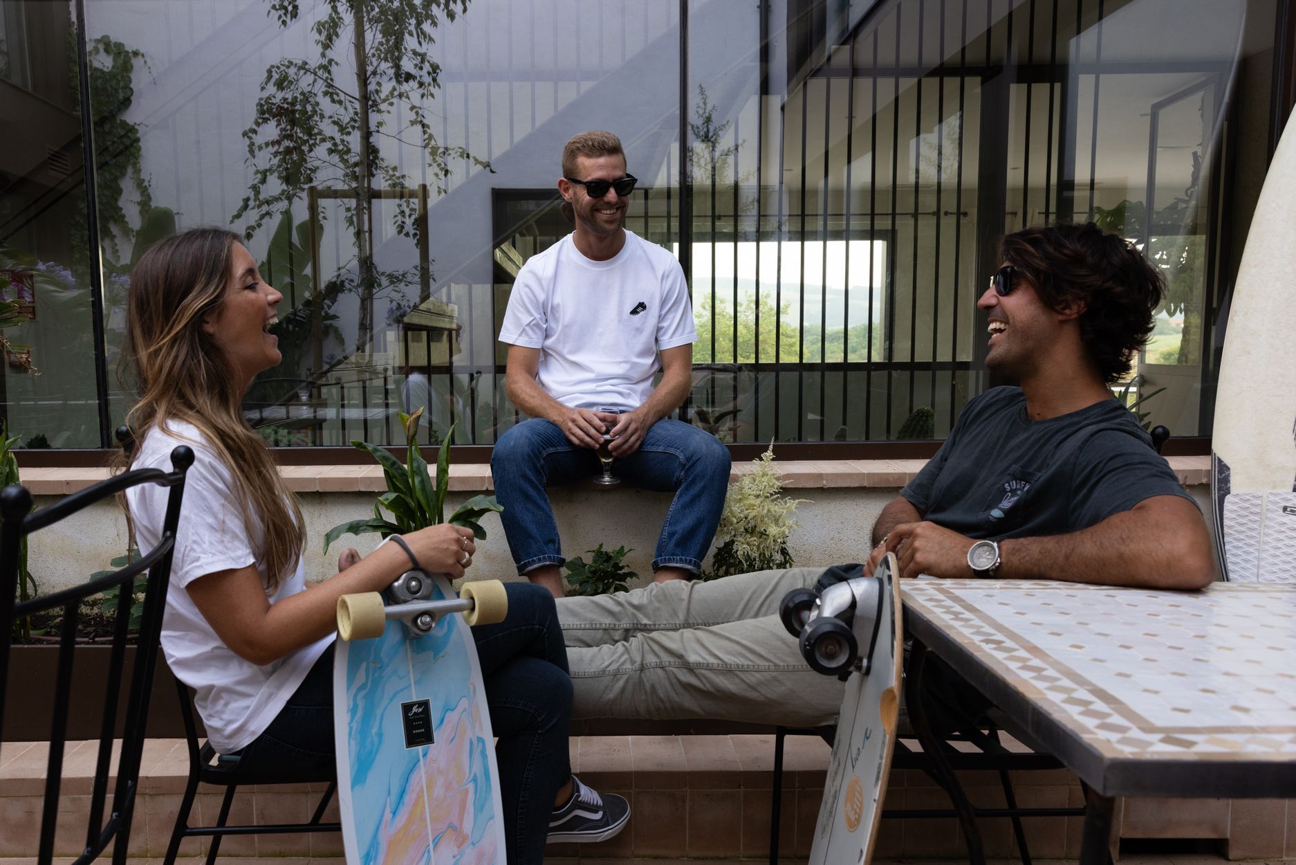 Tres personas charlando en una mesa de una cafetería al aire libre, con bancos y un edificio moderno al fondo.