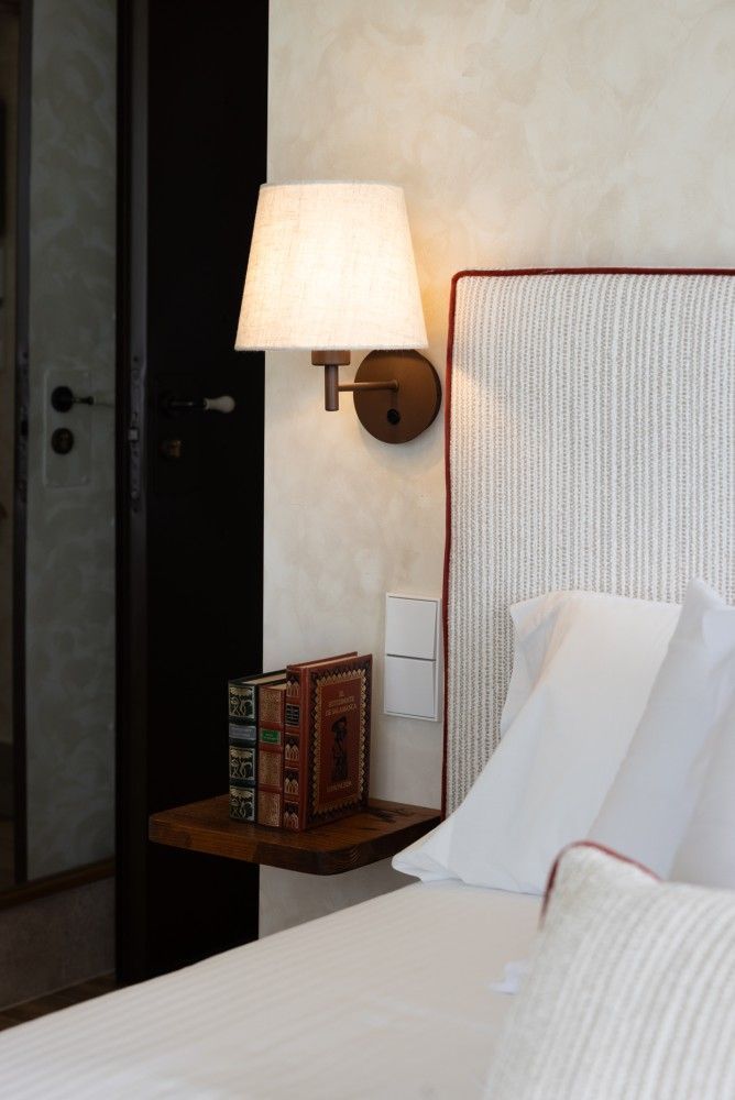 Una cama bien hecha con sábanas blancas, junto a una lámpara de pared y una pequeña mesita de noche de madera.