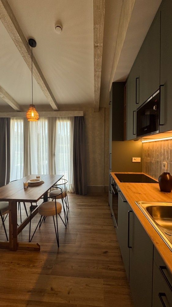 Cocina y comedor modernos con armarios verdes, suelos de madera cálida y una mesa junto a una ventana con cortinas.