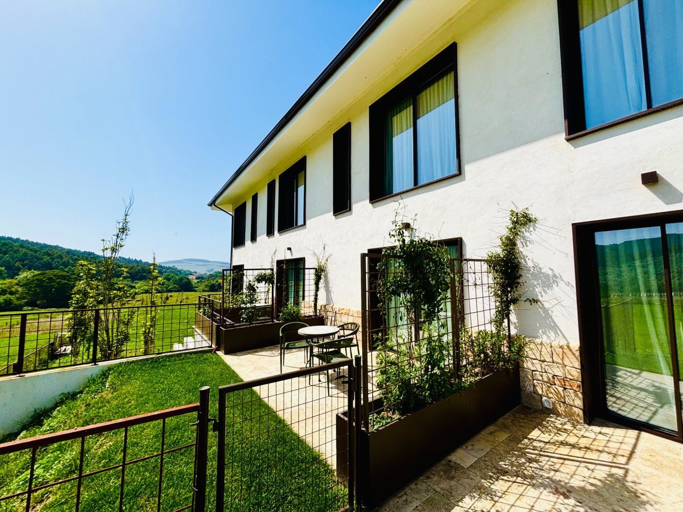 Casa blanca moderna con grandes ventanales, terraza y un césped verde con vistas a las colinas.