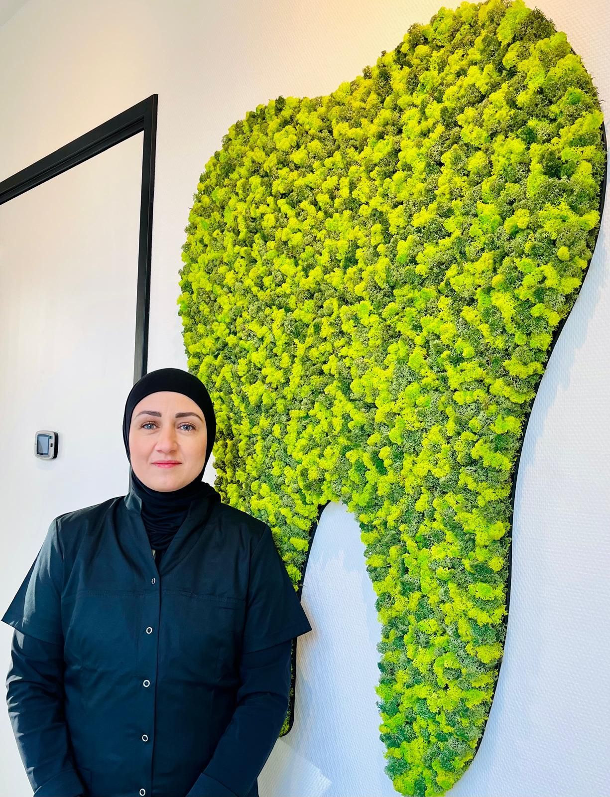 Vrouw in zwart uniform, staande naast een grote, groene, mosachtige, tandvormige wanddecoratie.