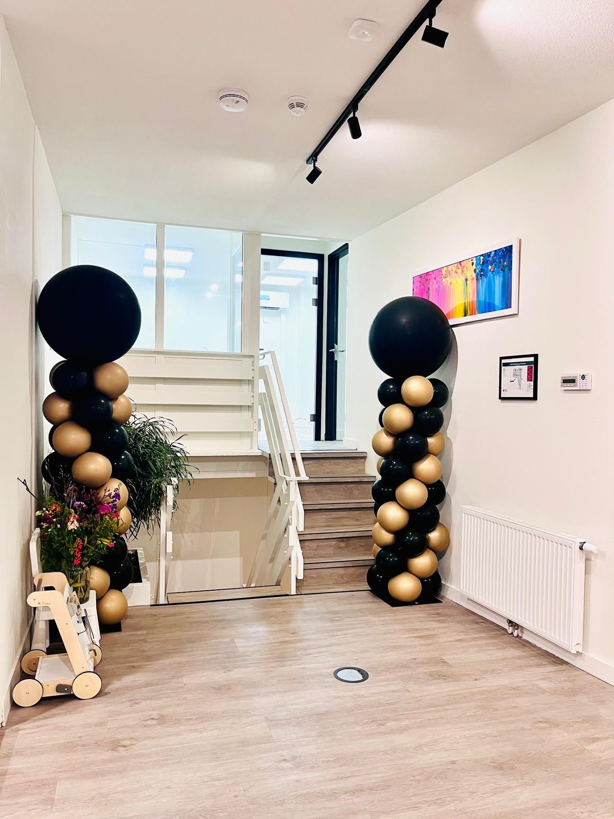 Twee ballonzuilen met gouden en zwarte ballonnen flankeren een hal met een trap en een lichte houten vloer.
