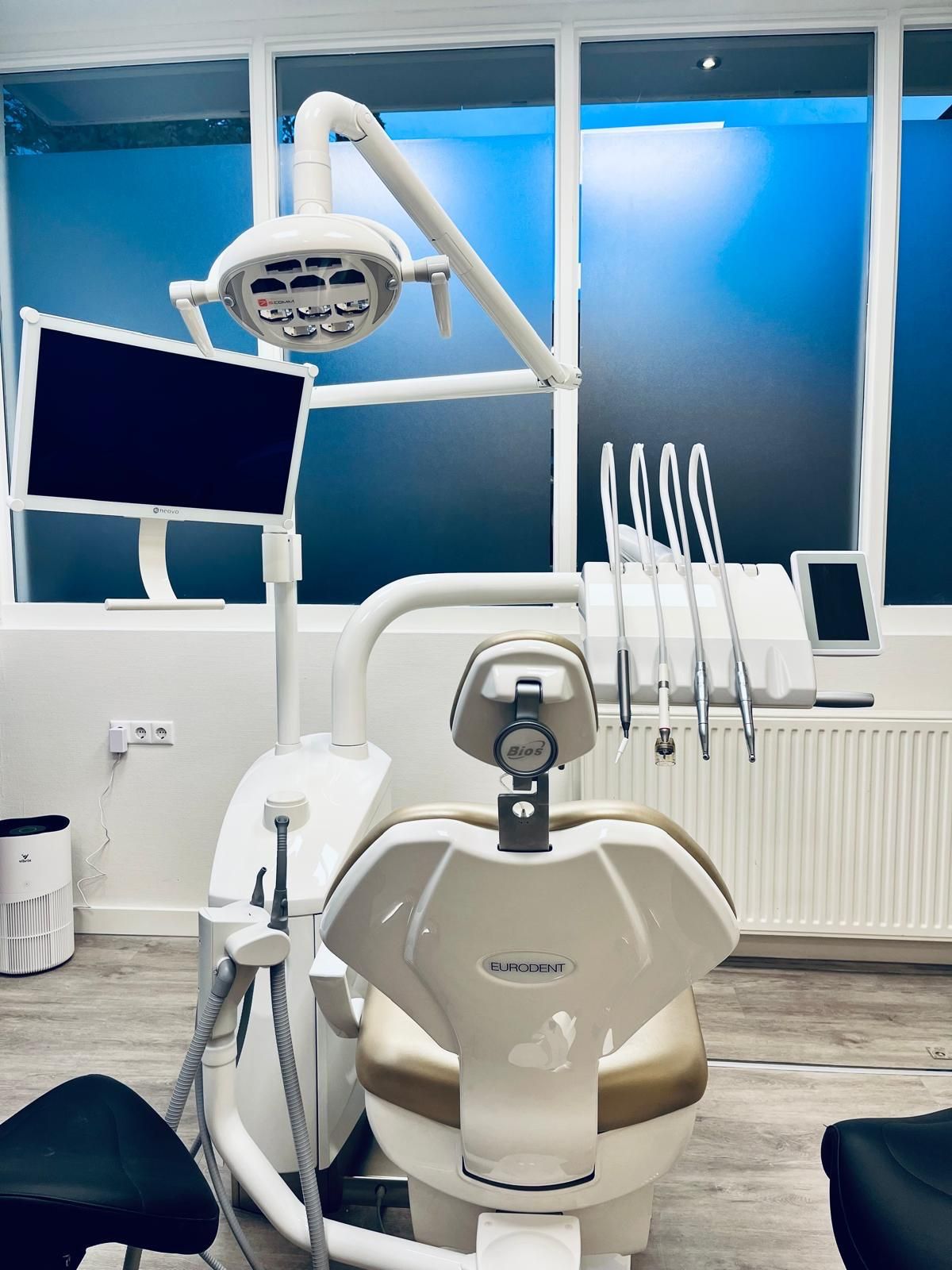 Tandartsstoel en -apparatuur in een lichte, moderne praktijk met een monitor en raam.