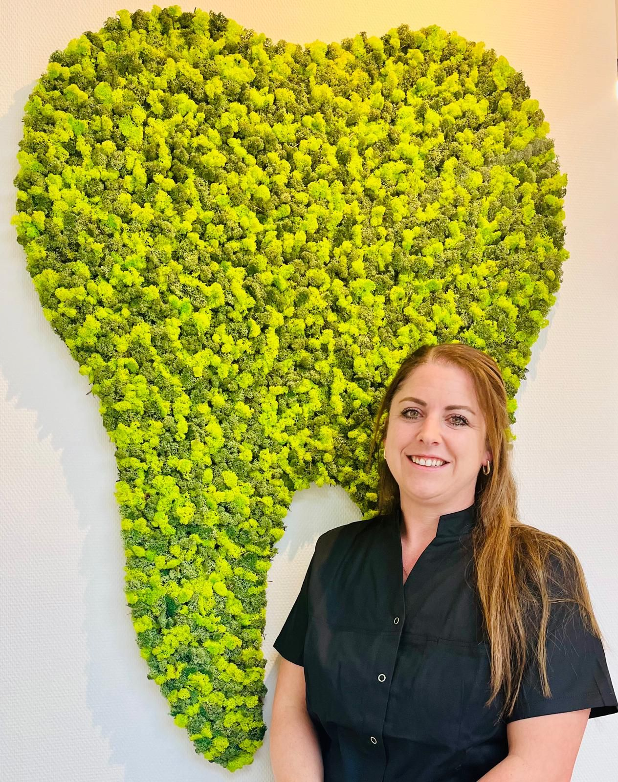 Een vrouw in een zwarte operatiekleding lacht voor een limoengroene, met mos bedekte tand op een witte muur.