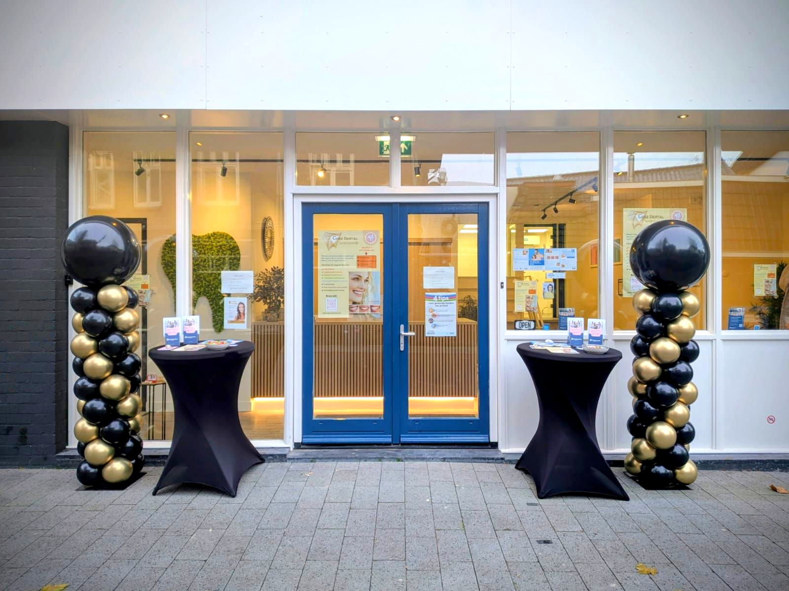 De ingang is versierd met ballonzuilen en tafels. Een blauwe deur, gouden en zwarte ballonnen en etalages.