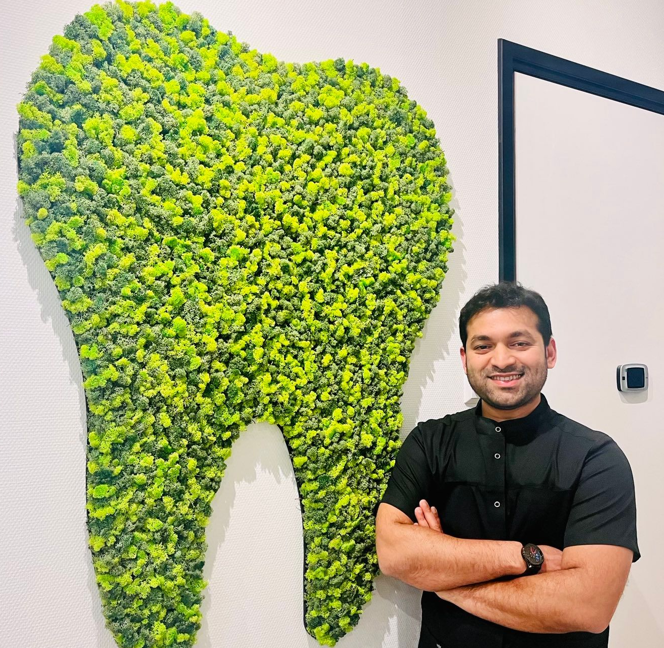 Een man die lacht, met zijn armen over elkaar, staat naast een grote, groene, tandvormige muurdecoratie.