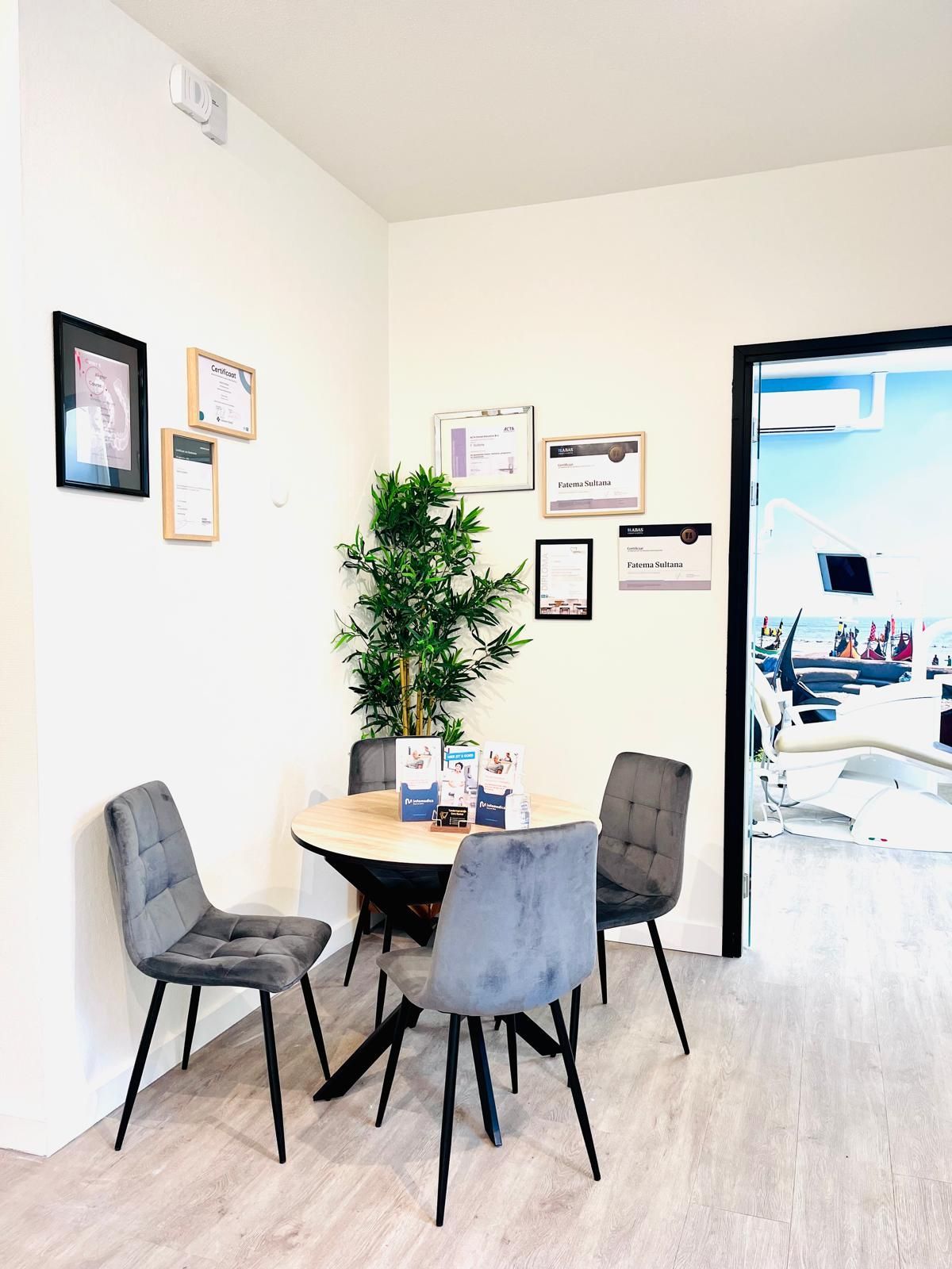 Een kleine wachtruimte met een ronde tafel en drie grijze stoelen. Een plant en ingelijste documenten sieren de muur.