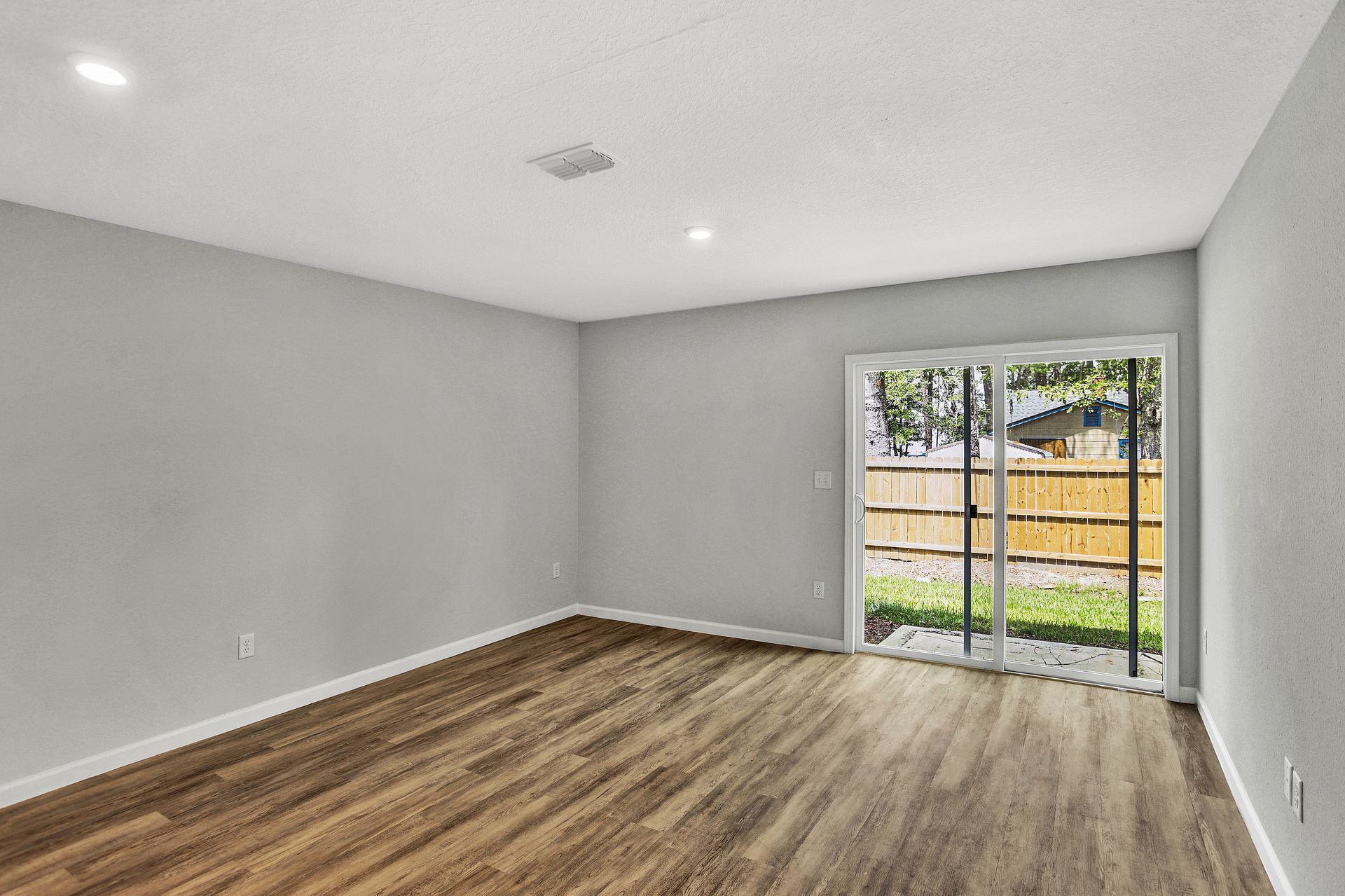 Empty room with gray walls, wood flooring, and a sliding glass door to the backyard.