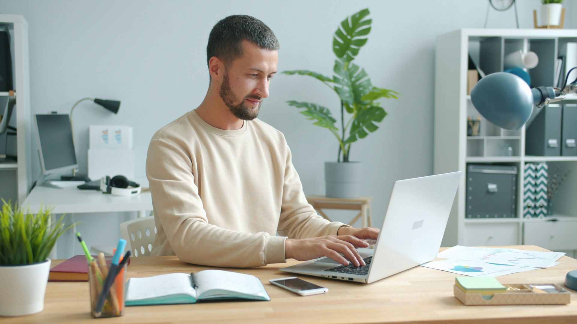 Man typing on a laptop at a desk in a home office with a plant, phone, and notebooks.