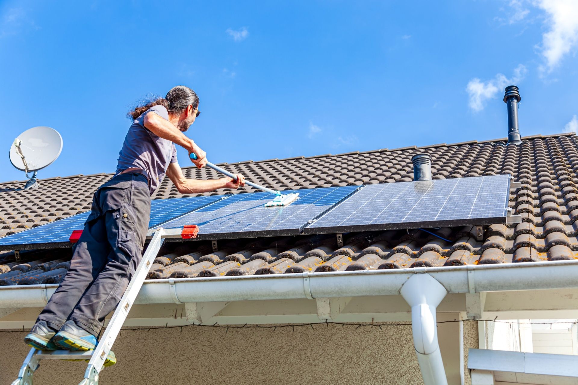A man is standing on a ladder installing solar panels on the roof of a house.