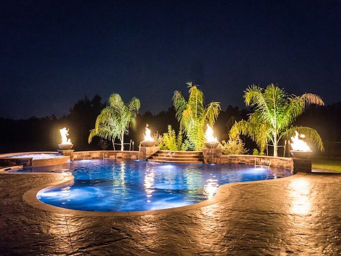 A large swimming pool is lit up at night with palm trees in the background.