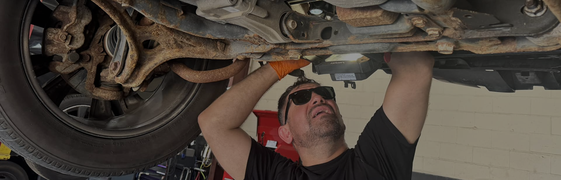 Mechanic working under a car, wearing sunglasses and gloves. Rusty undercarriage, tires in view. | Auto Check Elite