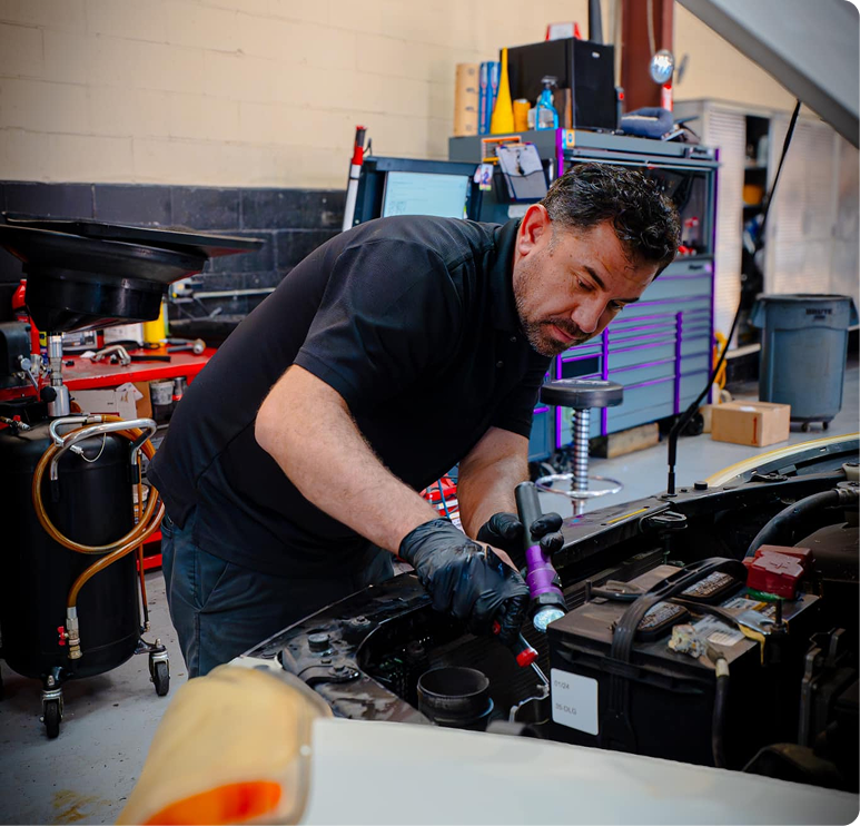 Mechanic working on car engine, using a flashlight. Workshop setting with tools and equipment. | Auto Check Elite