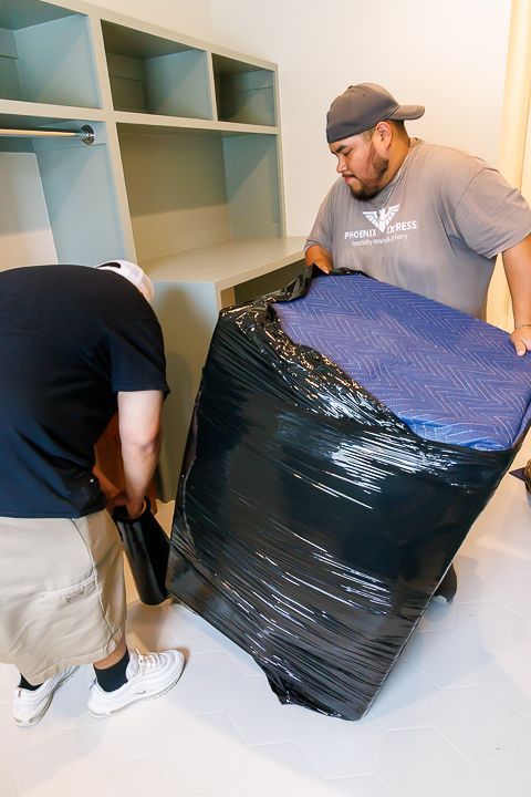 Two people carrying a large, wrapped object in a room with built-in shelves.