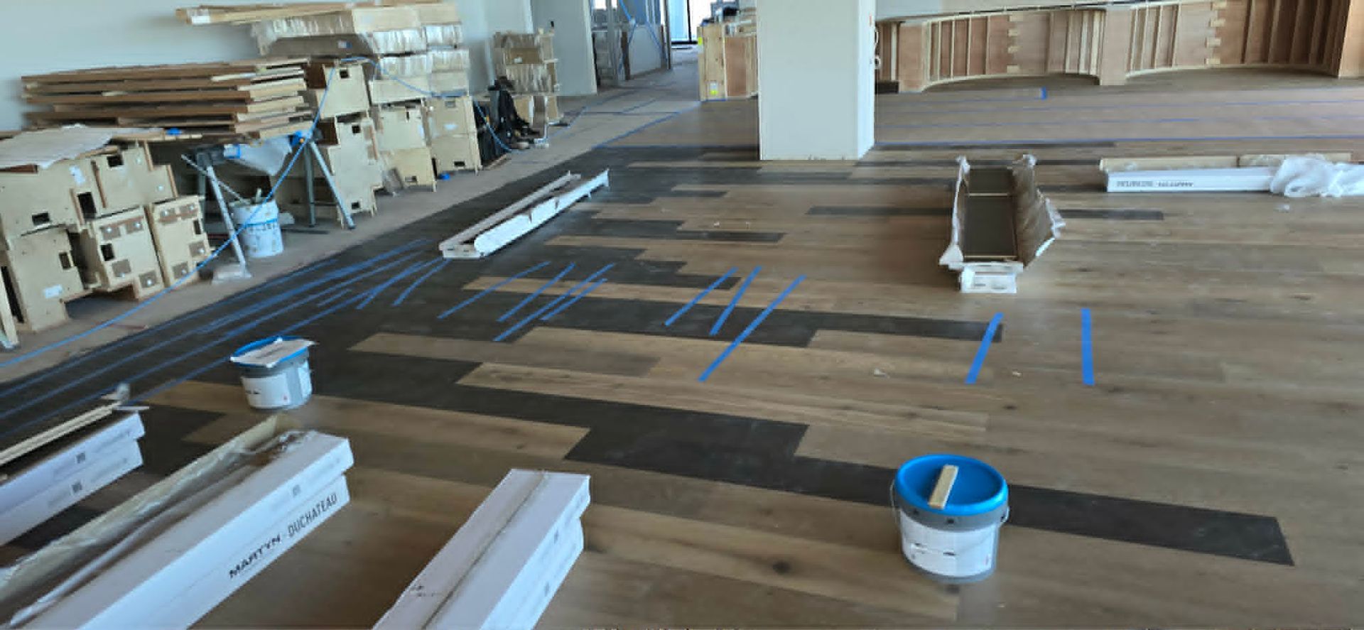 Wide workshop with wood planks, lumber stacks, and blue tape markings on the floor during construction.