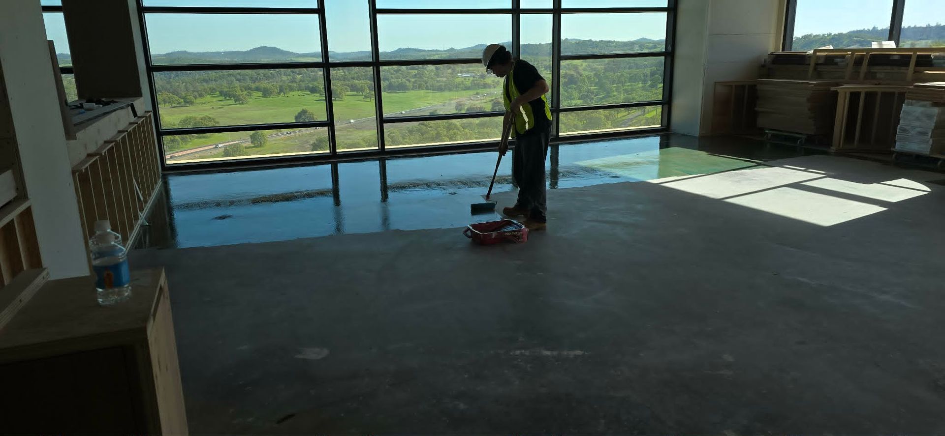 Person cleaning a sunlit room with a mop, standing near large windows overlooking a grassy field.