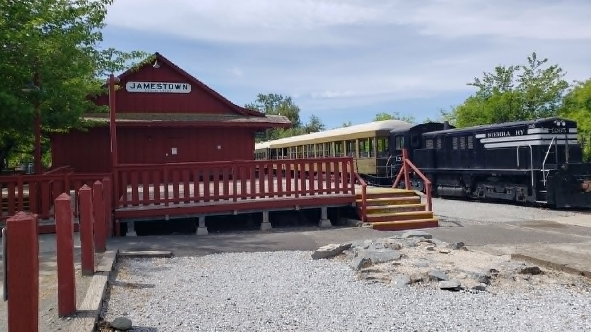 Red wooden train depot with porch, next to a black locomotive on tracks under a cloudy sky.