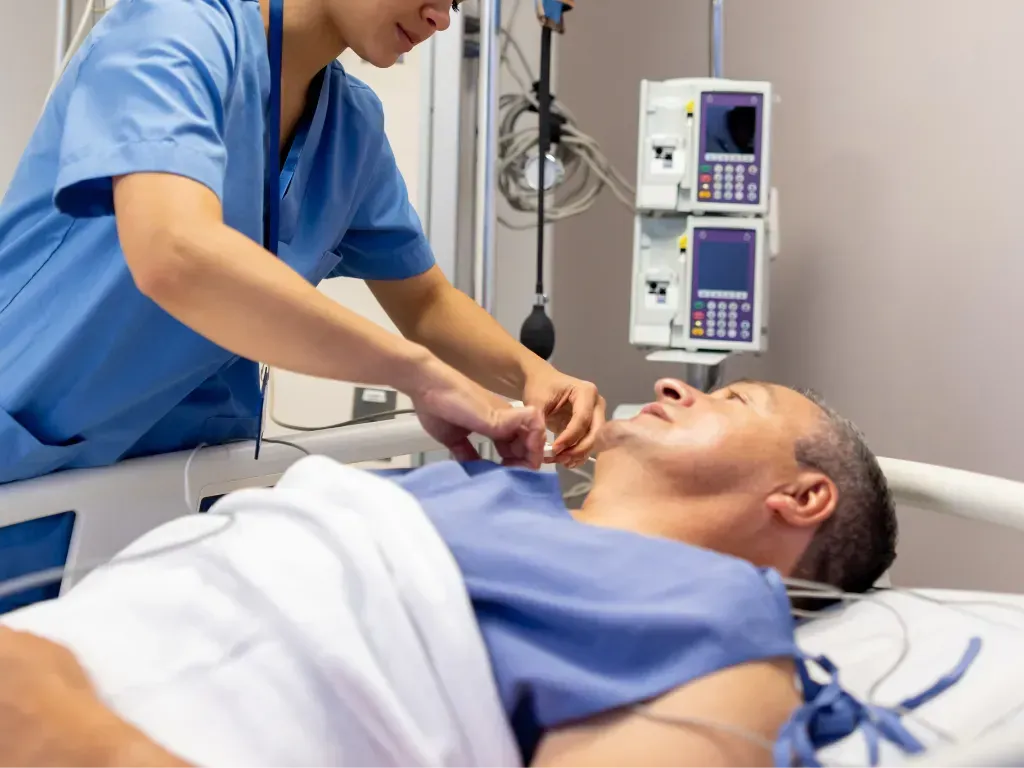 A healthcare professional in a blue uniform attends to a patient lying in a hospital bed with medical equipment nearby.
