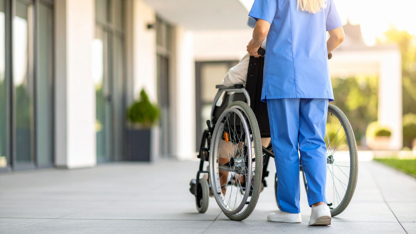 A caregiver in blue scrubs pushes a person in a wheelchair along an outdoor building walkway.