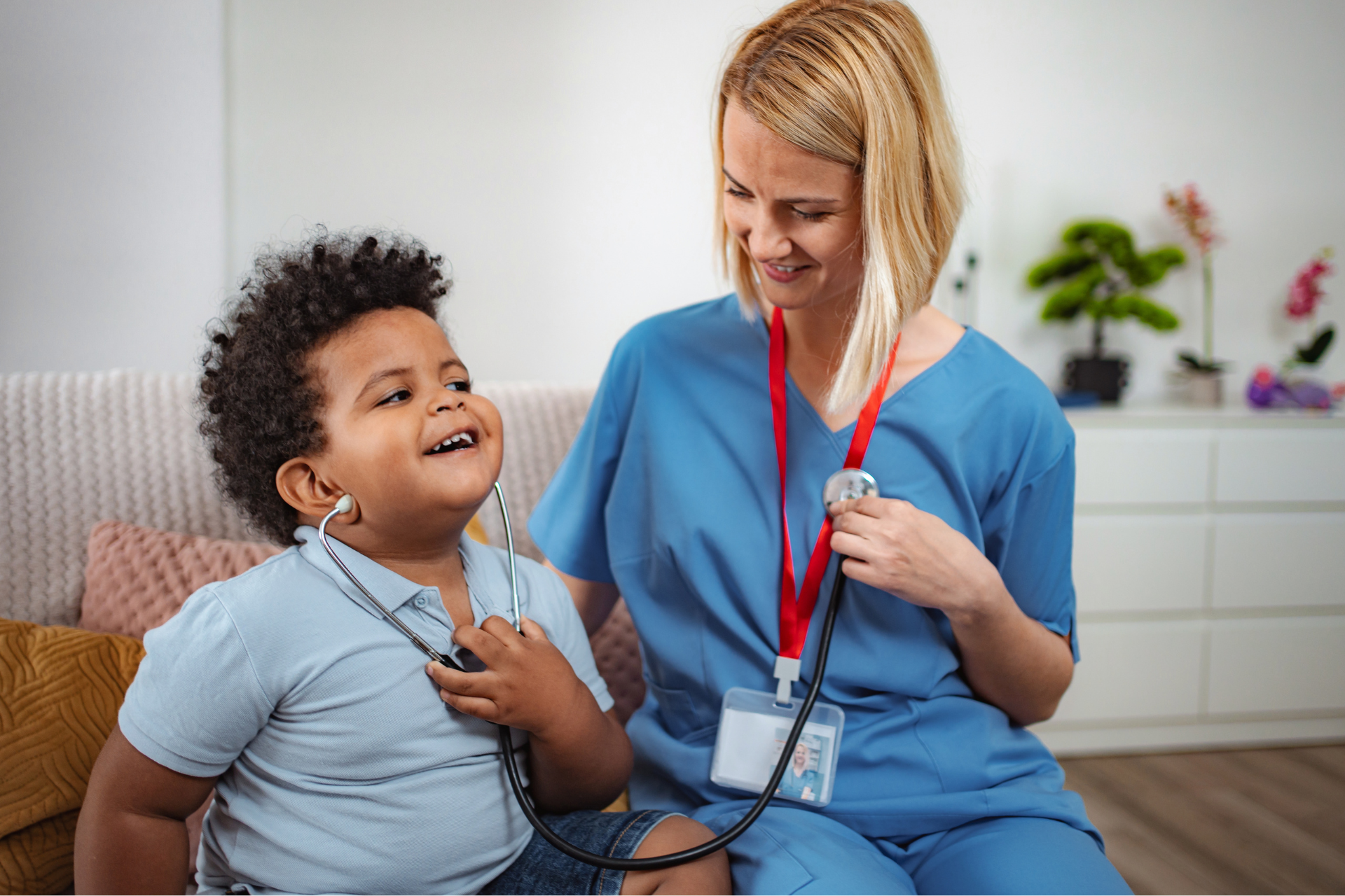 A healthcare provider in blue scrubs sitting on a sofa while a young child uses a stethoscope to listen to their own heart.