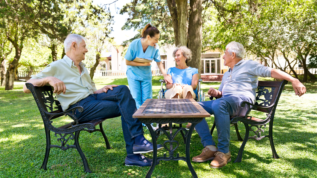A nurse interacts with two seated individuals at a small outdoor table in a park-like setting.