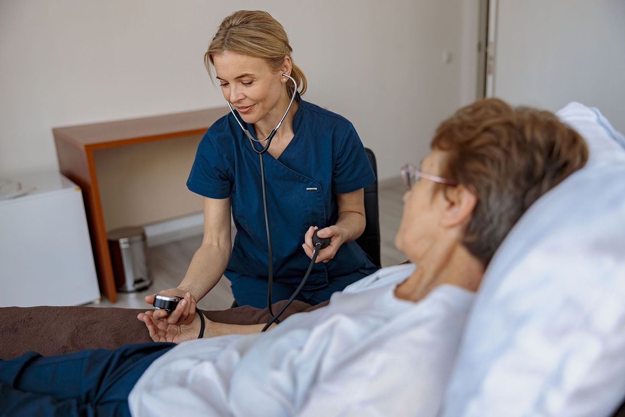 A healthcare worker in blue scrubs uses a pulse oximeter on a patient’s finger in a medical room.
