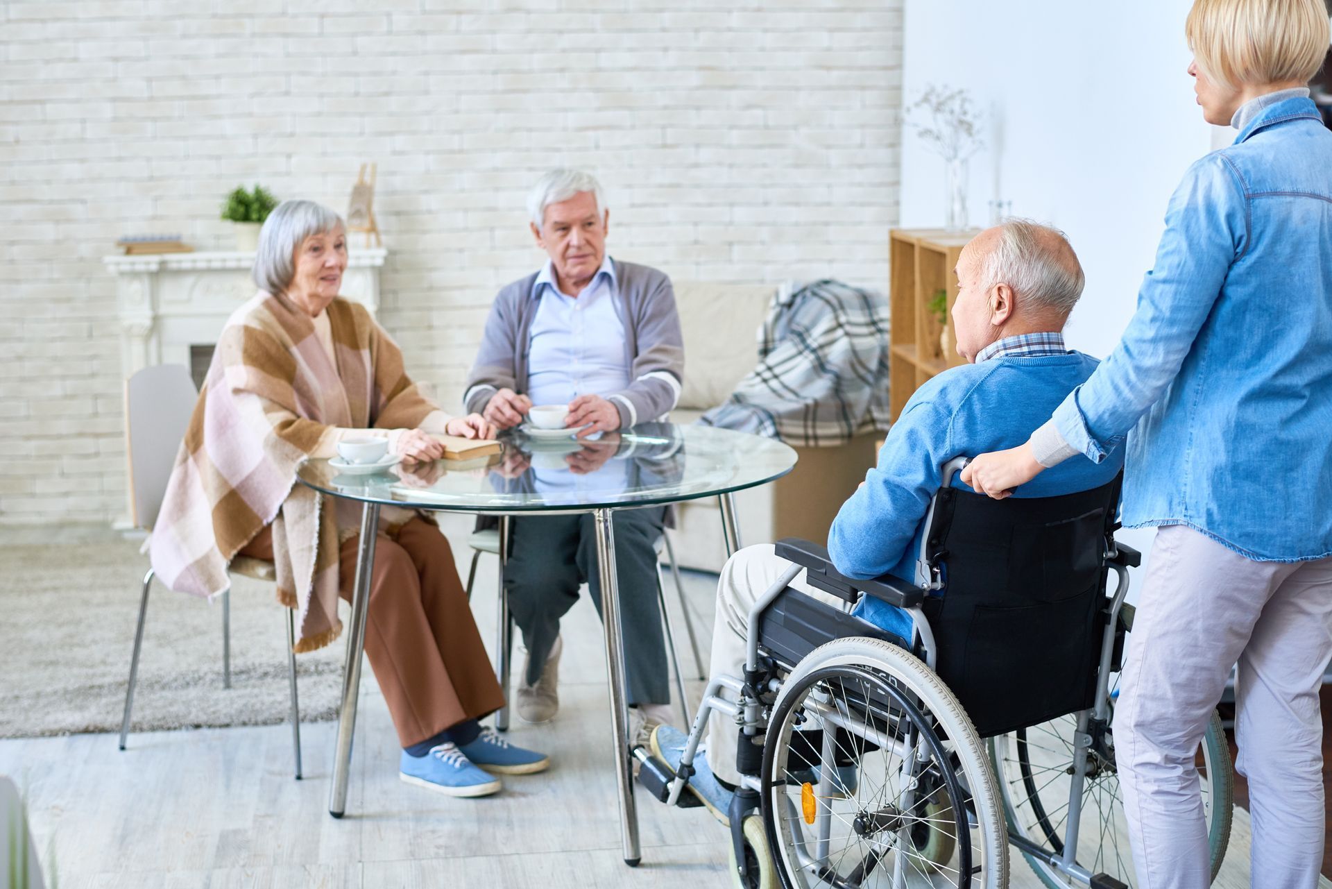 Three people sit at a glass table in a bright room, while a fourth person stands by a wheelchair in the foreground.