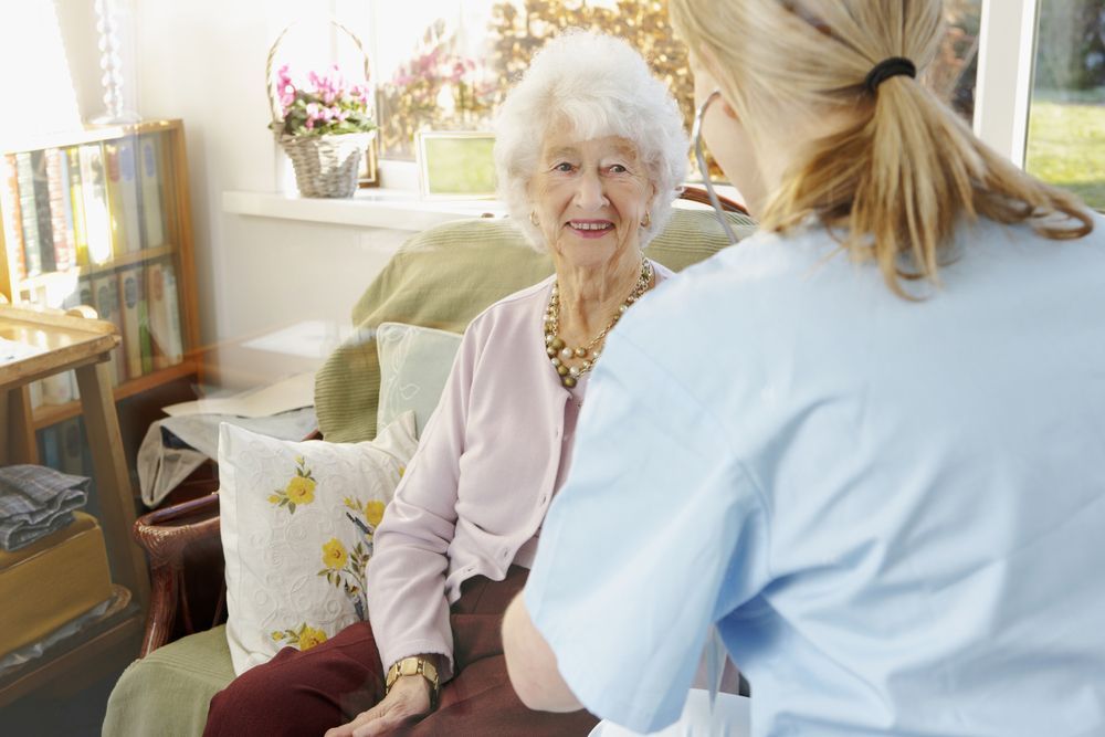 A person in scrubs using a stethoscope to examine a patient sitting in a chair in a bright, home-like setting.