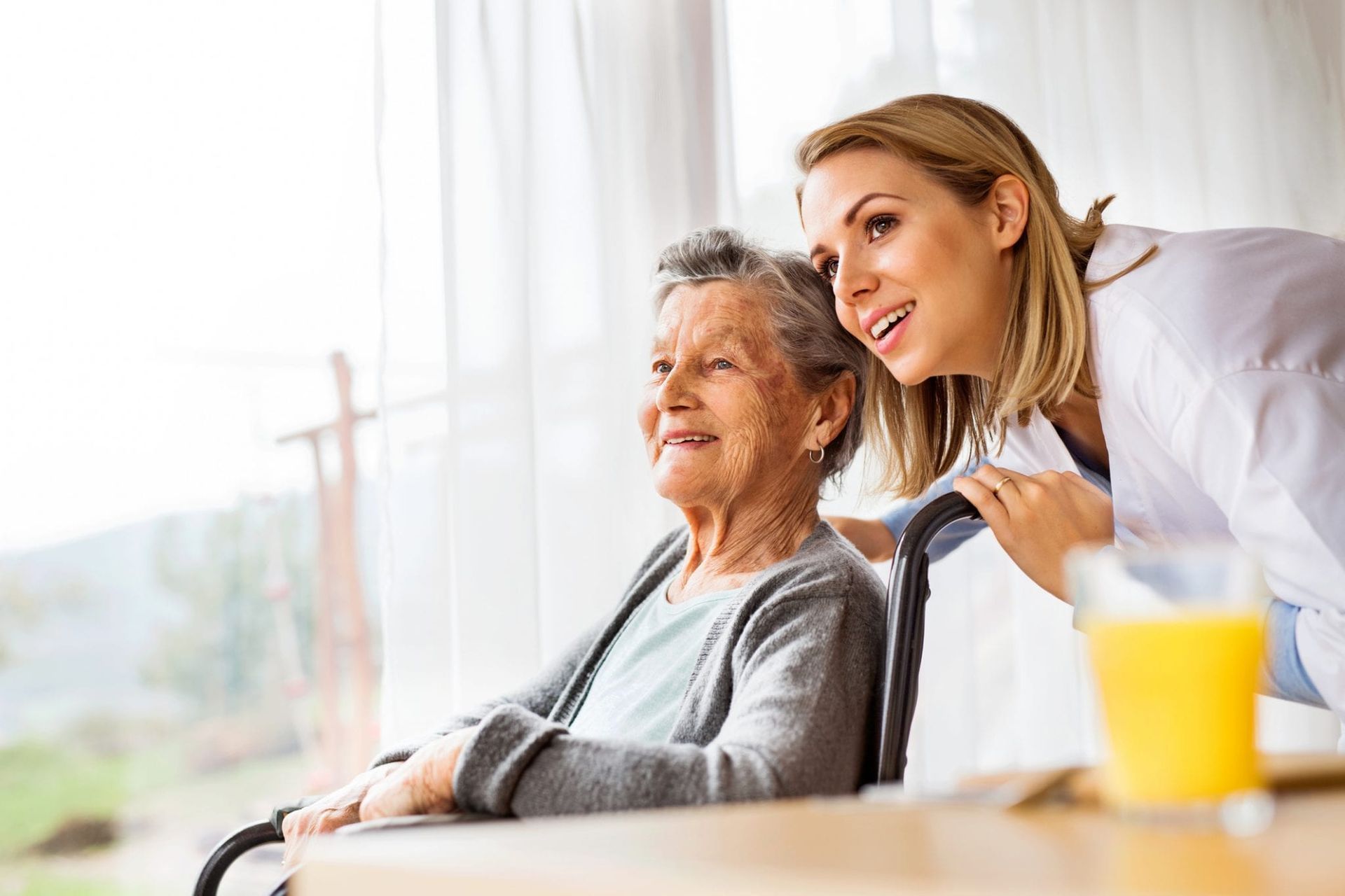 A caregiver in a white coat leans toward an individual in a wheelchair as they look out a window together.