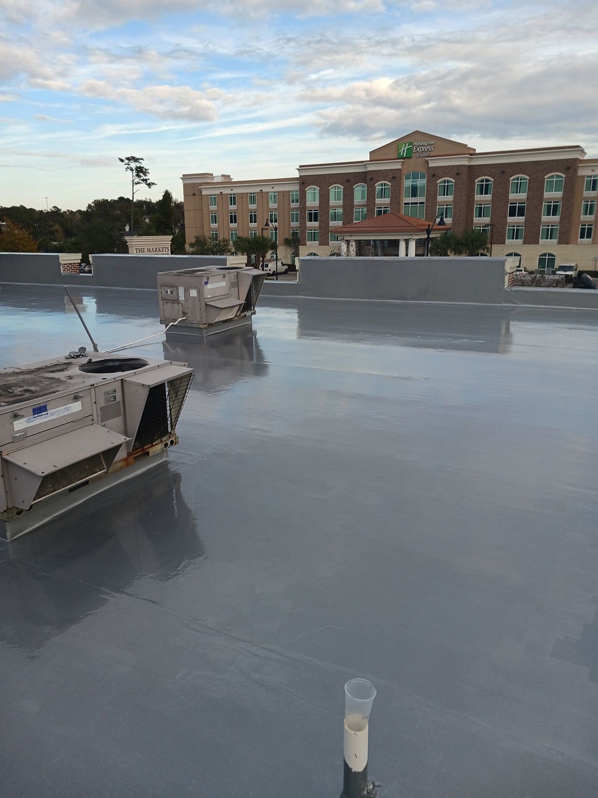 Flat gray commercial roof with HVAC units, and a building in the background under a cloudy sky.